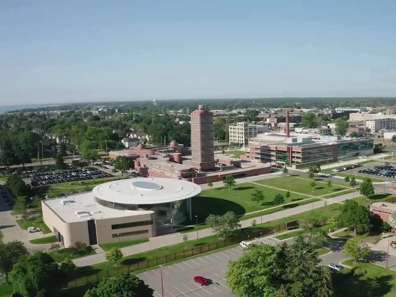 A large circular building with a flat roof is surrounded by trees and parking lots in a university campus setting, under a clear blue sky.