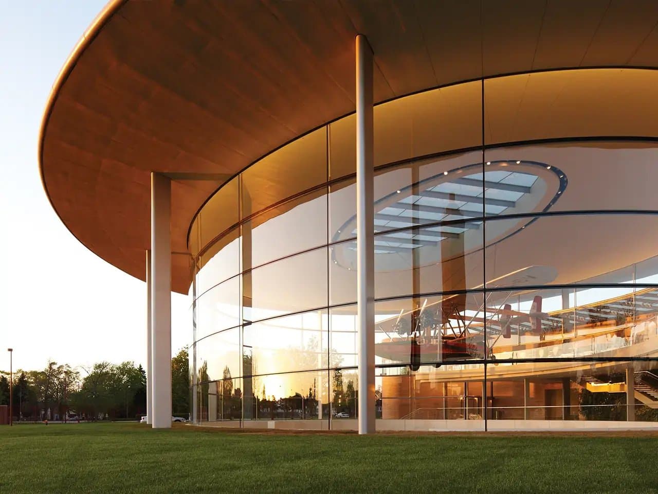 A modern, glass-walled building with a large circular roof reflects the sunset. Inside, a red aircraft is visible. The structure is surrounded by a grassy area and trees.