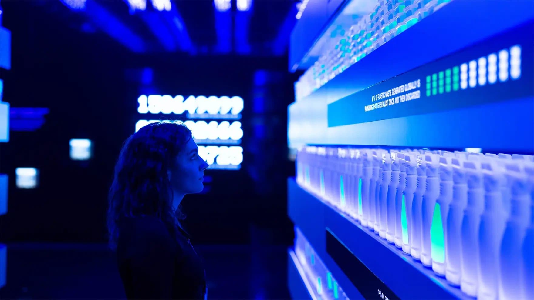 A person observes a display of illuminated bottles against glowing blue signage reading "40% OF PLASTIC WASTE... DISPOSED." The scene takes place in a futuristic, darkly lit setting.