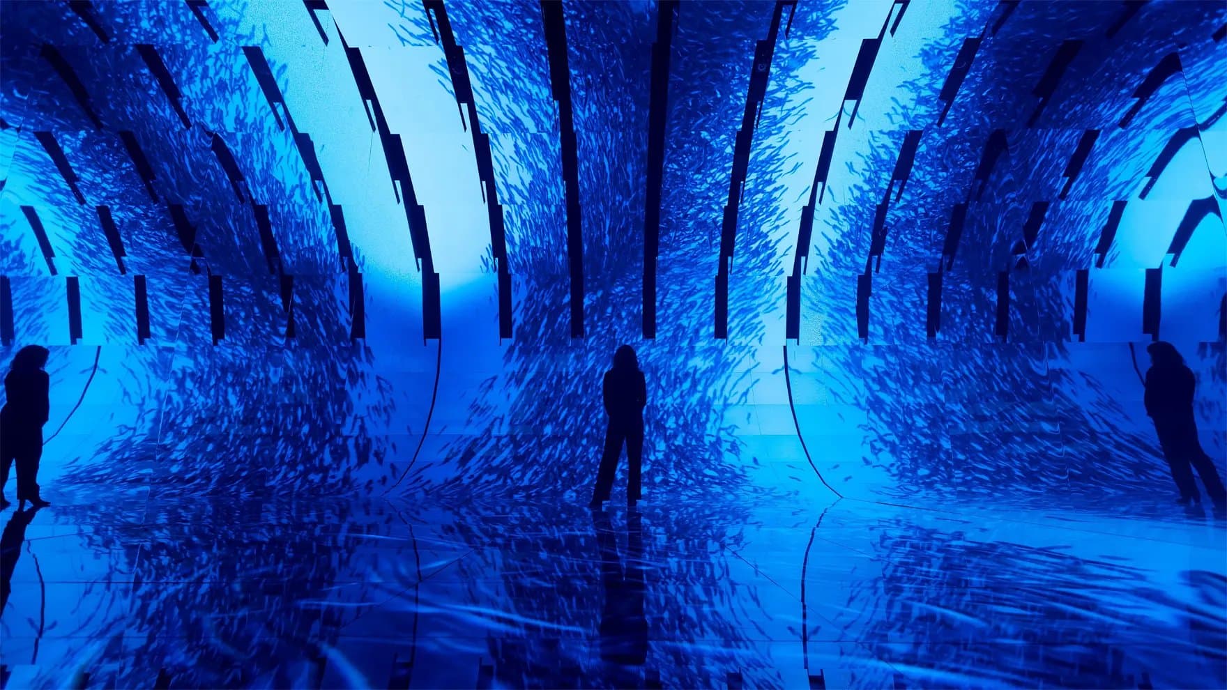 Silhouetted figures stand within an immersive digital art installation; a flowing, textured blue display surrounds them, creating an oceanic atmosphere inside a dark, reflective space.