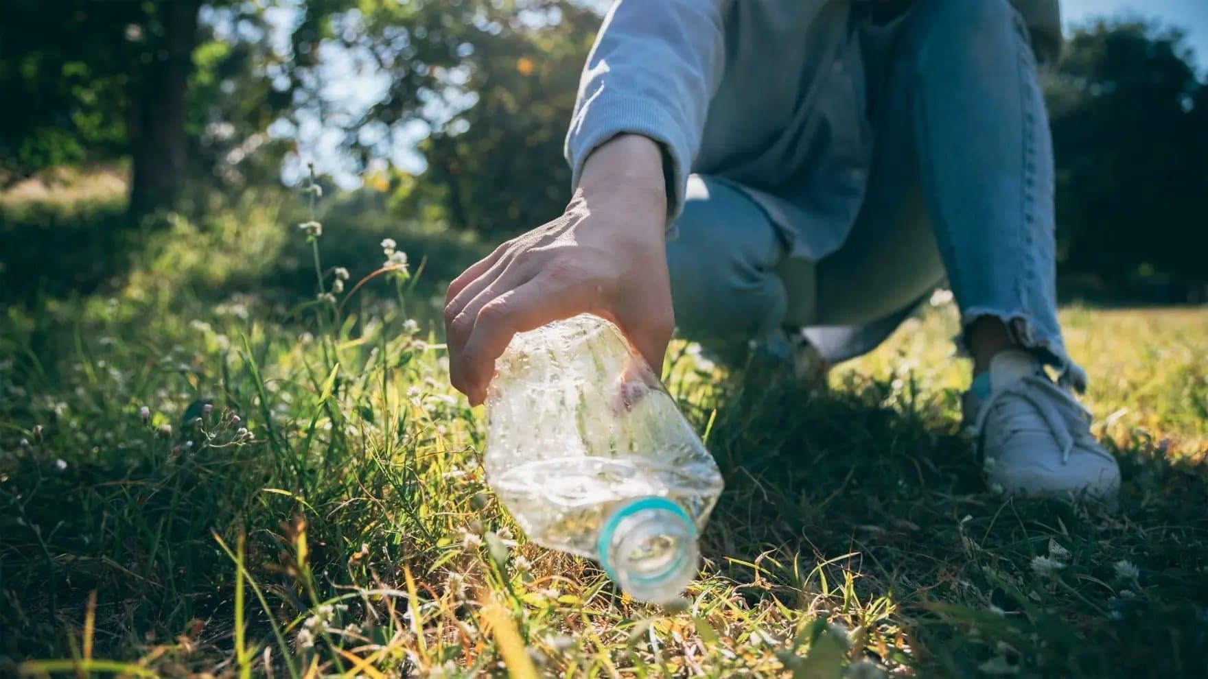 A hand picks up a crumpled plastic bottle in a sunlit grassy field, suggesting environmental cleanup or recycling efforts.