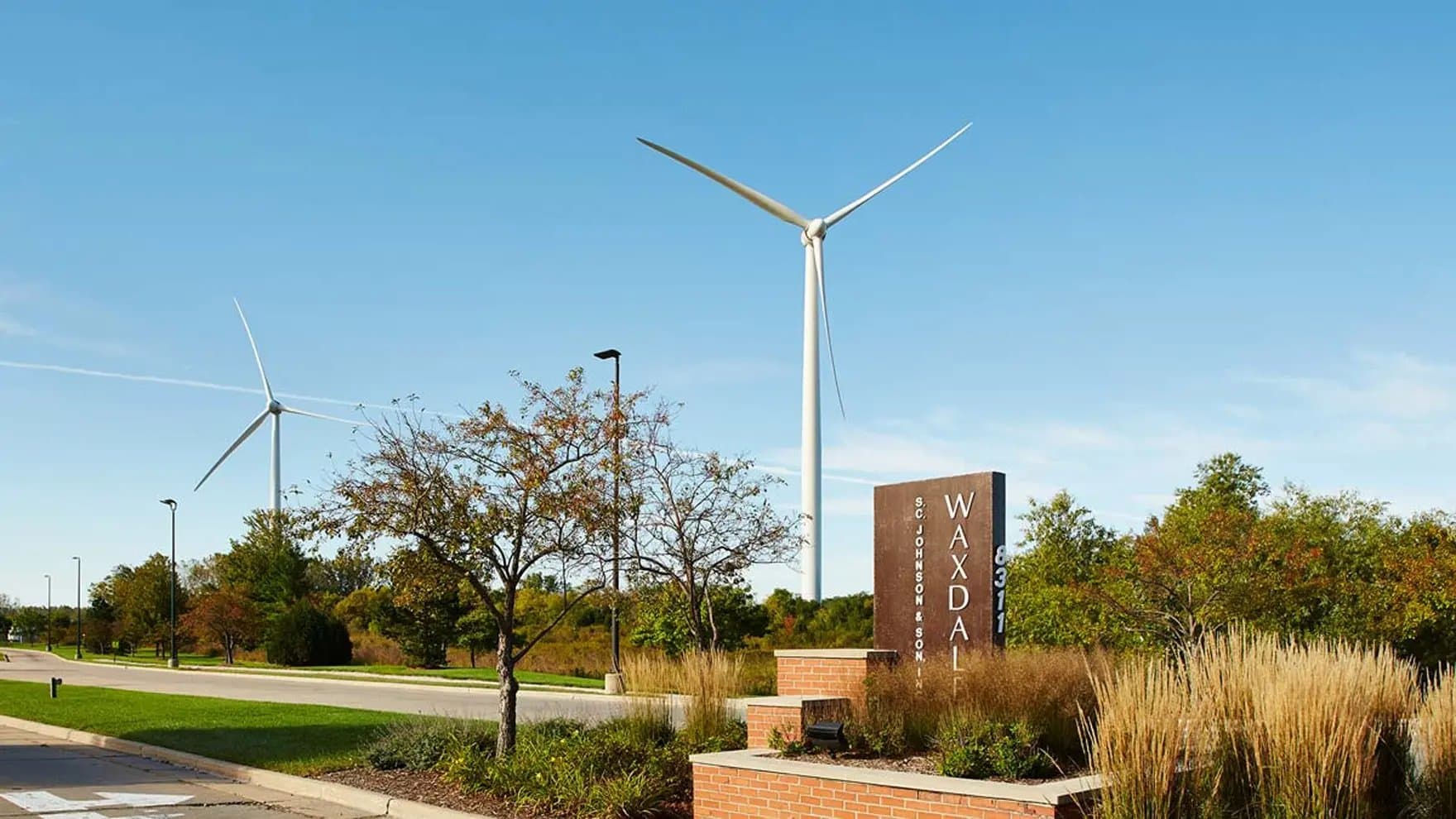 A wind turbine spins under a clear sky near a roadside. The foreground features a sign reading "S.C. Johnson & Son, Inc. Waxdale 8311" amidst grass and trees.