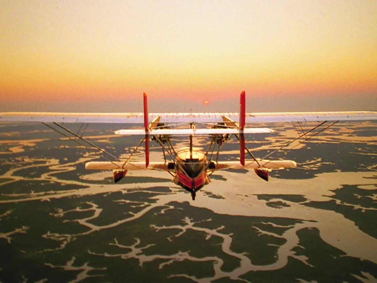 A biplane flies over a winding river landscape at sunset, with a vibrant sky blending orange and purple hues, silhouetting the intricate water patterns below.