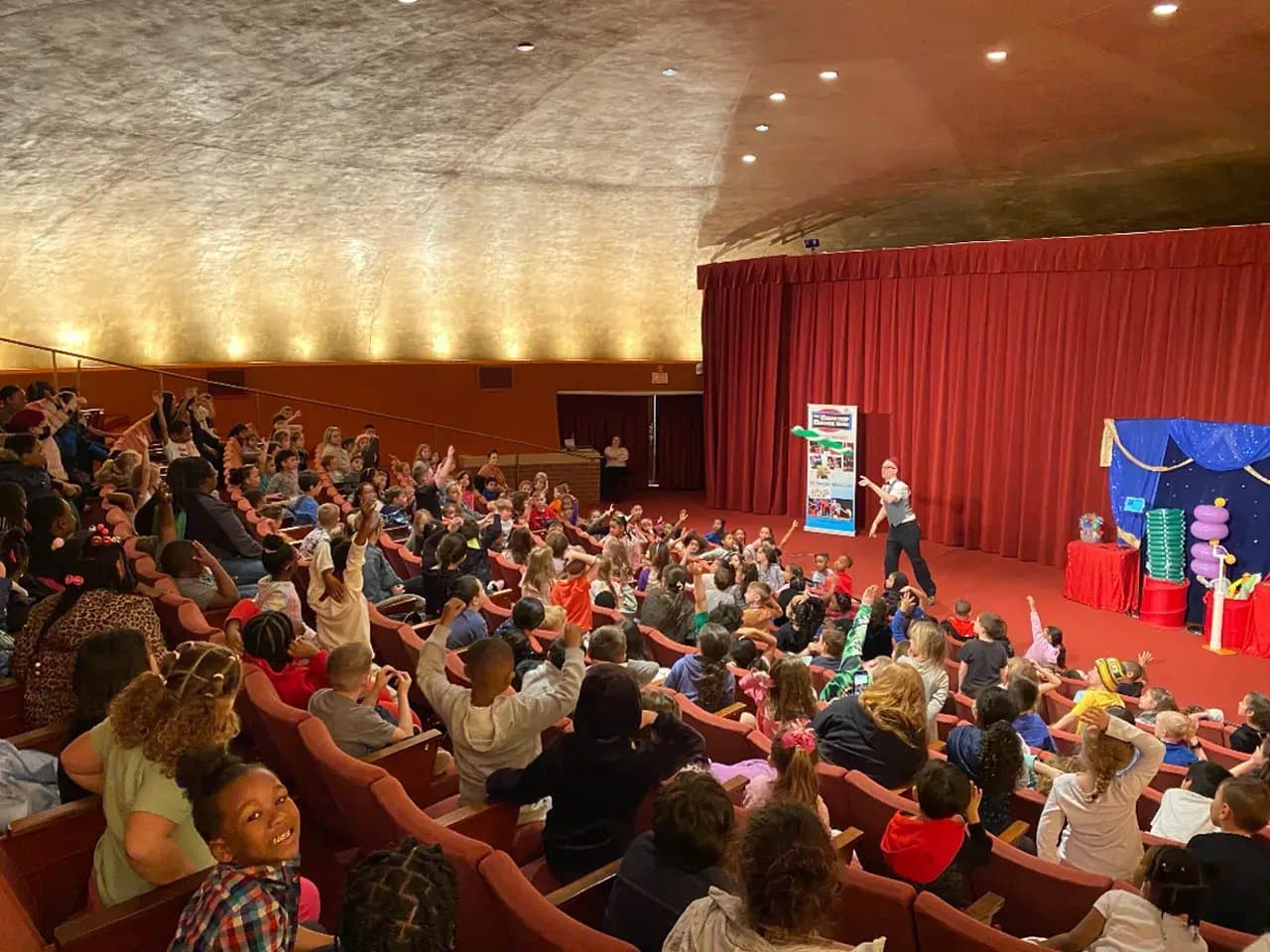 A performer entertains a large audience of children seated in a theater. The room is warmly lit, with red curtains and stage props, creating a lively, engaging atmosphere.