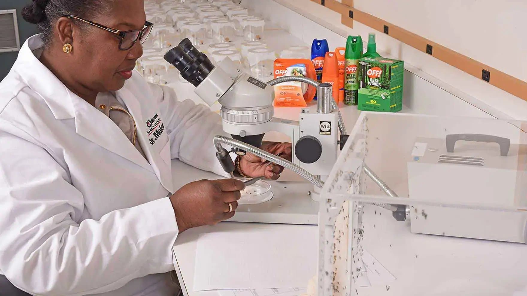 Scientist examines samples under a microscope in a lab setting. In the background, various OFF! insect repellent products are displayed. She wears a white coat labeled "Dr. M. Meek."