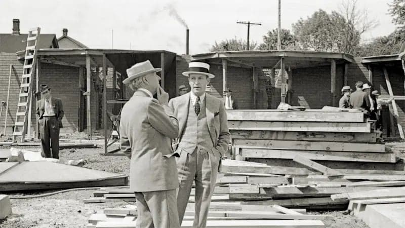 Two men in suits converse, standing amid scattered wooden planks at a construction site. In the background, several other men are gathered, and buildings with smoke stacks are visible.