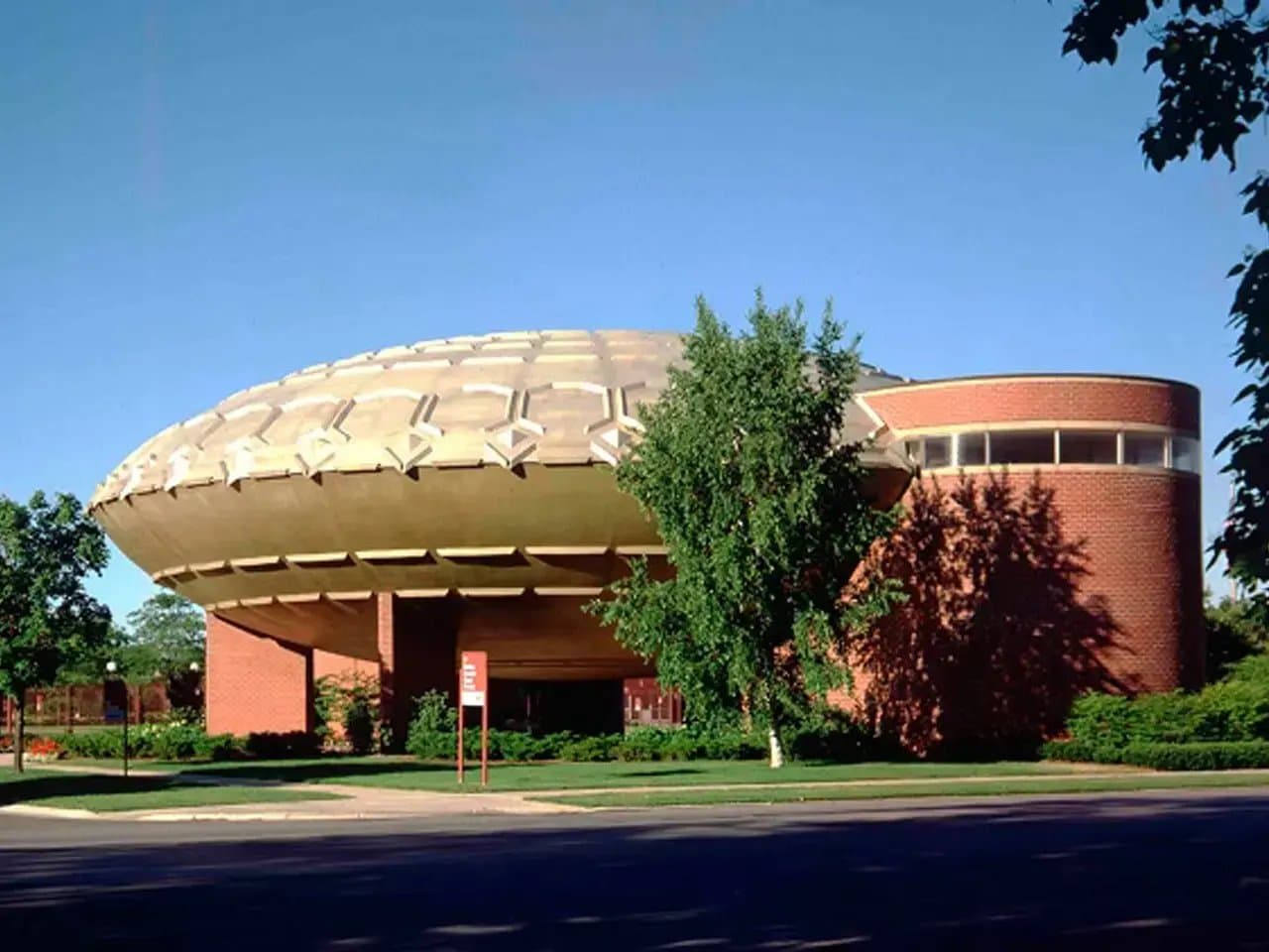 A uniquely shaped, round, dome-like building with a textured roof sits amidst trees and greenery, with a clear blue sky in the background.