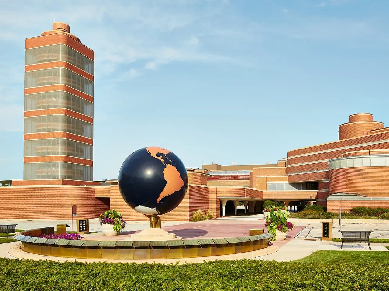 A large globe sculpture stands prominently, surrounded by neatly arranged flowers and benches, in front of a modern brick and glass building complex under a clear blue sky.