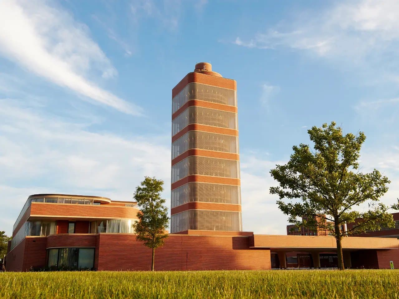 A tall, cylindrical brick tower stands amidst a modern building complex, surrounded by green lawns and trees under a blue sky, conveying a blend of architecture and nature.