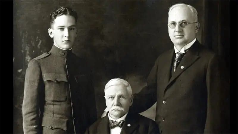 Three men pose in a formal setting; one stands in a military uniform, another is seated with a mustache, and the third stands in a suit with glasses, suggesting a historical context.