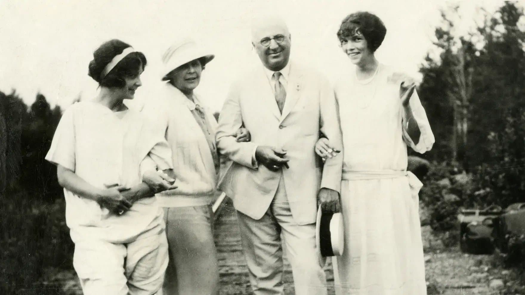 A group of four people, two women wearing hats and a man in a suit, stand closely together, smiling outdoors with trees in the background.