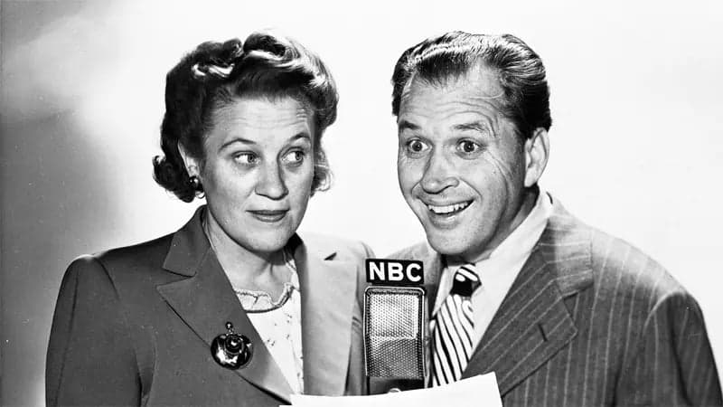 Two people stand together, speaking into an "NBC" microphone. The woman appears thoughtful, and the man looks enthusiastic. They are dressed in mid-20th-century formal attire, with a monochromatic backdrop.