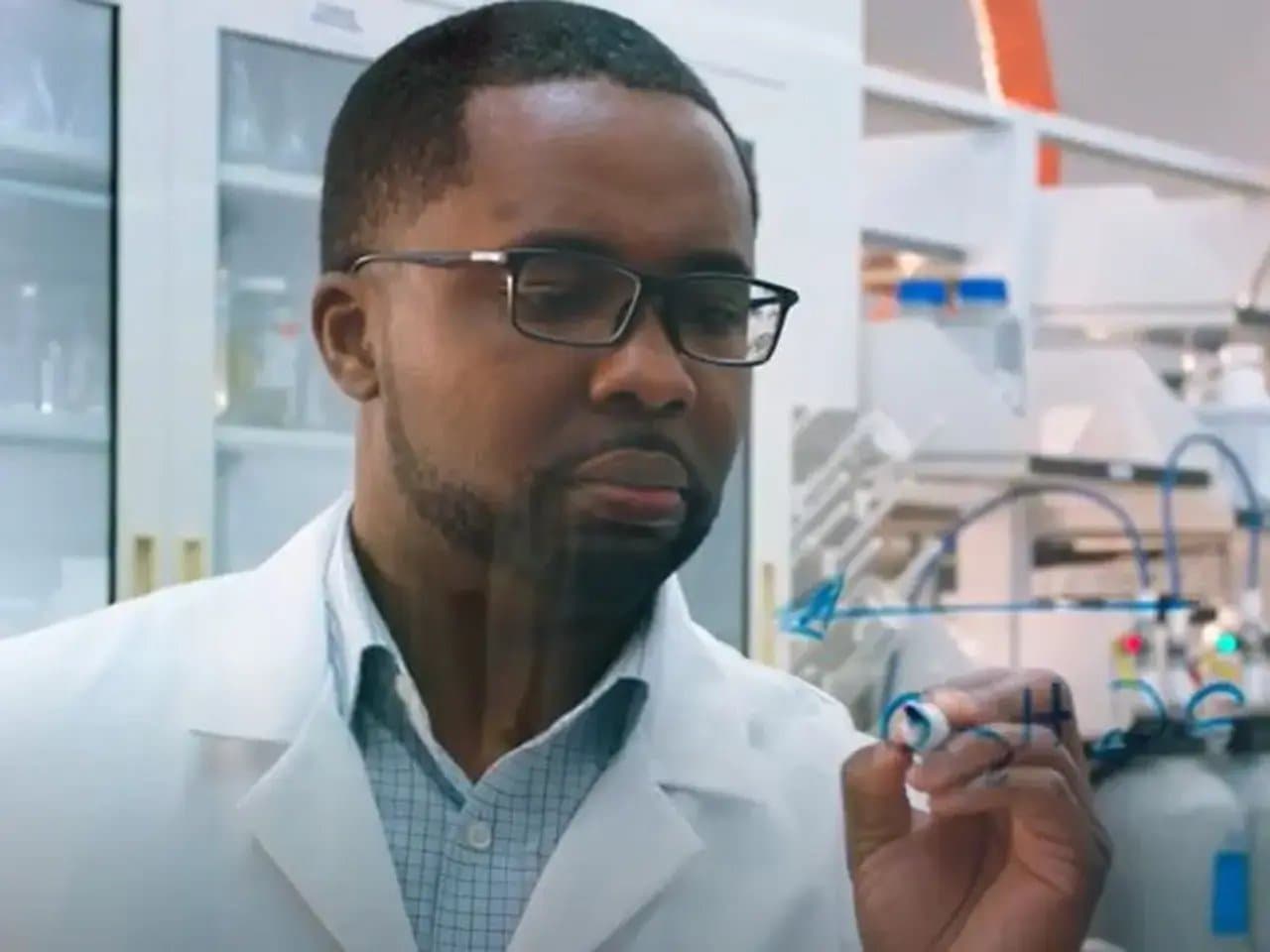 A scientist in a lab coat writes a chemical formula on a transparent board, surrounded by shelves with scientific equipment in a laboratory setting.