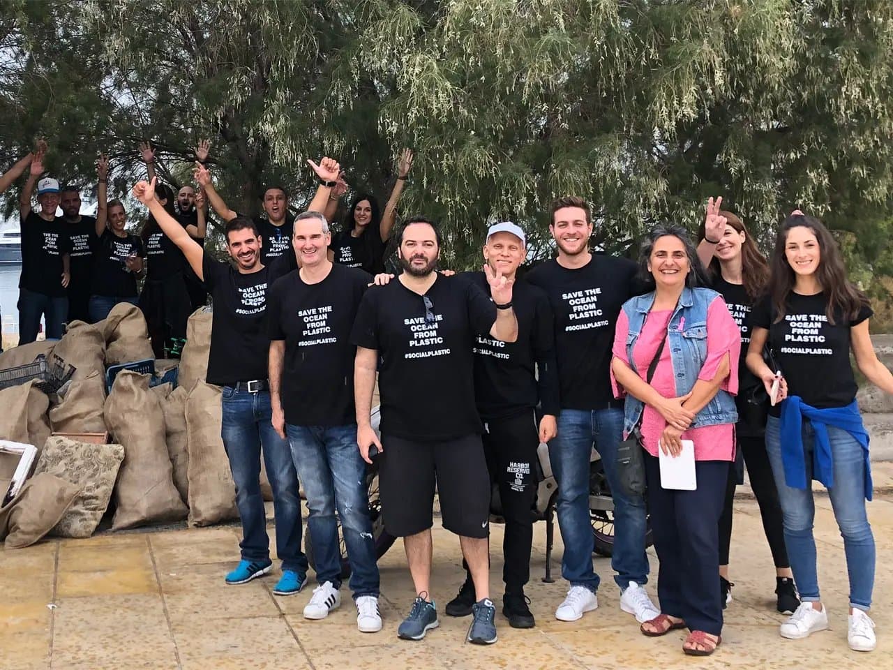 Group of people wearing "SAVE THE OCEAN FROM PLASTIC #SOCIALPLASTIC" shirts, posing happily. Baskets and bags are piled behind them, and trees form the backdrop, suggesting a cleanup event.