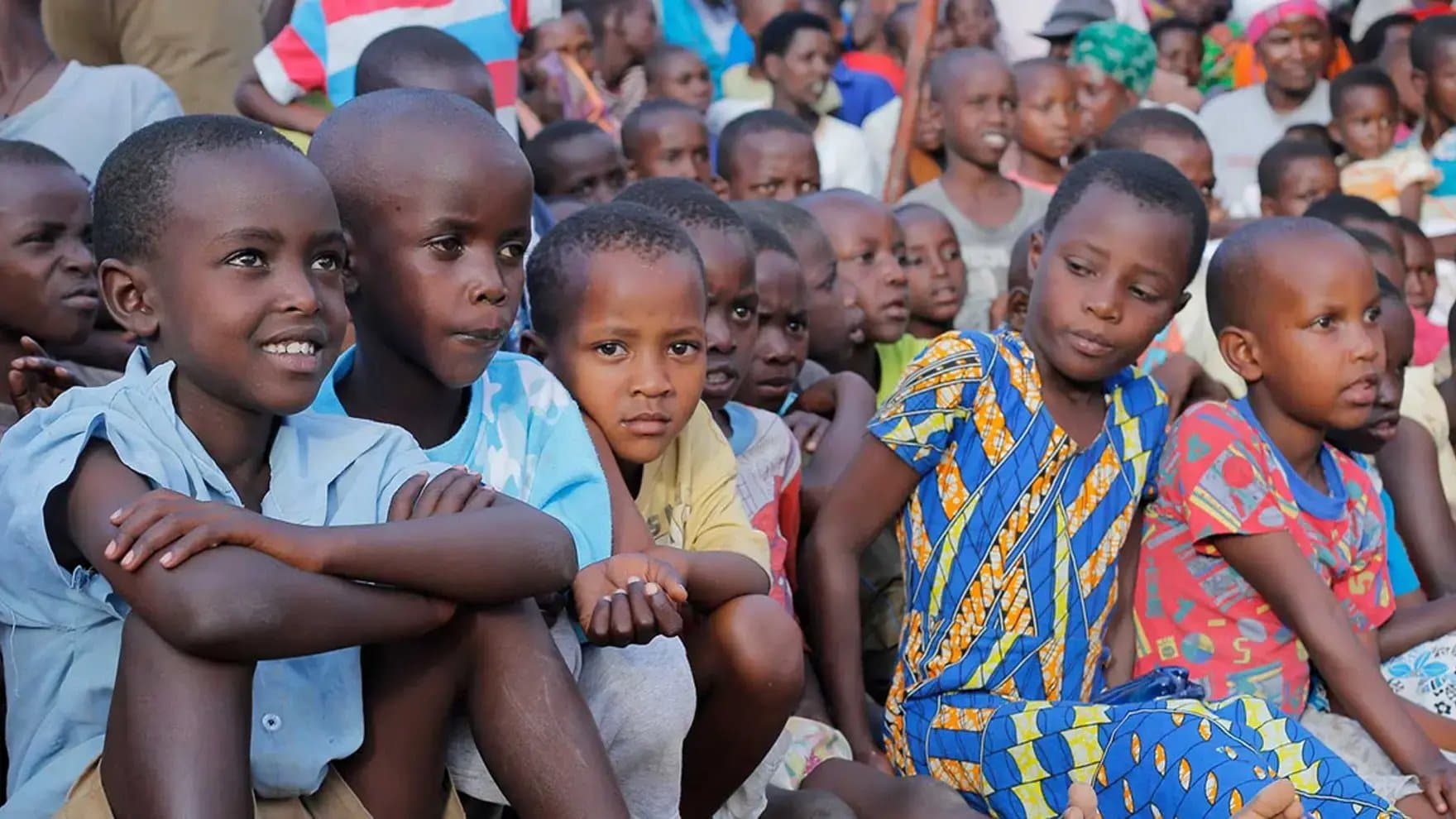 Children sit closely together, attentively watching something, in an outdoor gathering. They wear colorful clothing, surrounded by a crowd of people in a lively, communal setting.