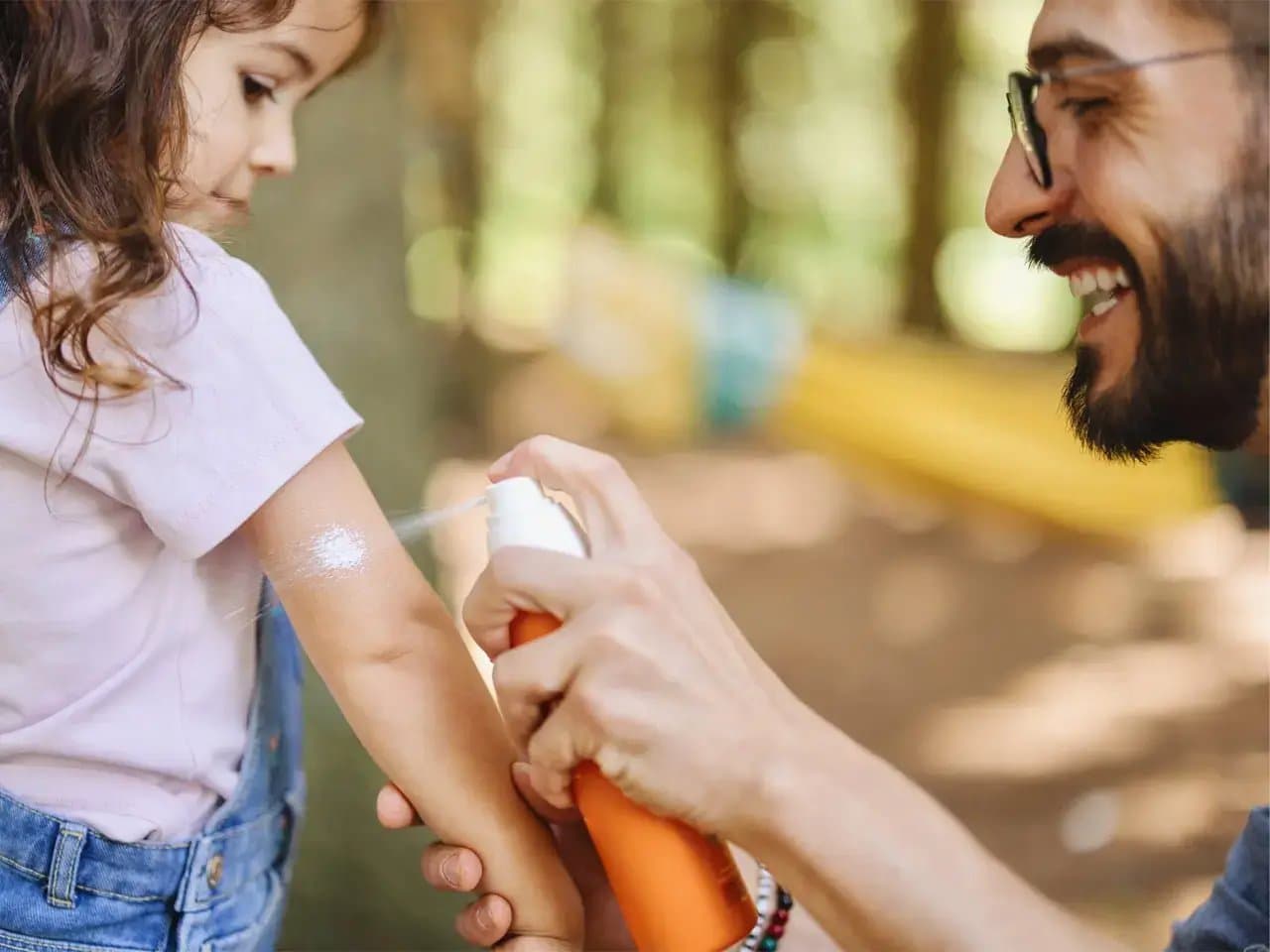 Child receives spray sunscreen on arm from an adult holding an orange bottle, outdoors in a wooded area with dappled sunlight.