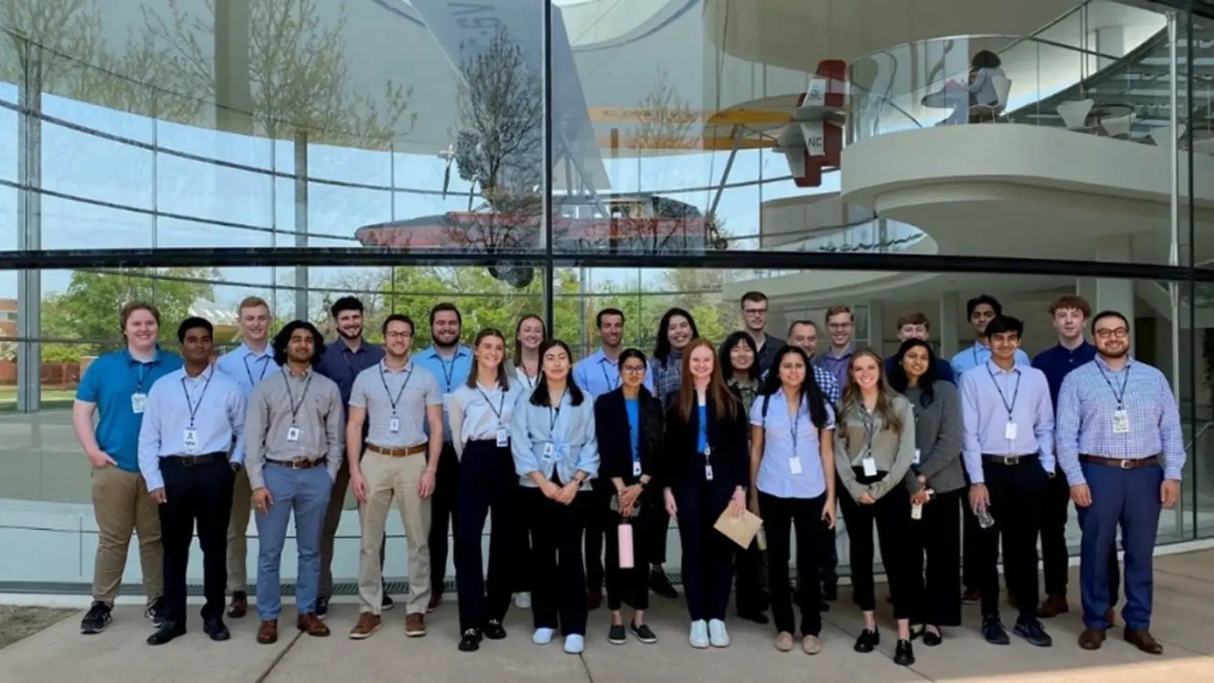 A group of people stand together, smiling, outside a modern glass building. The background reflects a tree and an interior balcony with a small aircraft display.