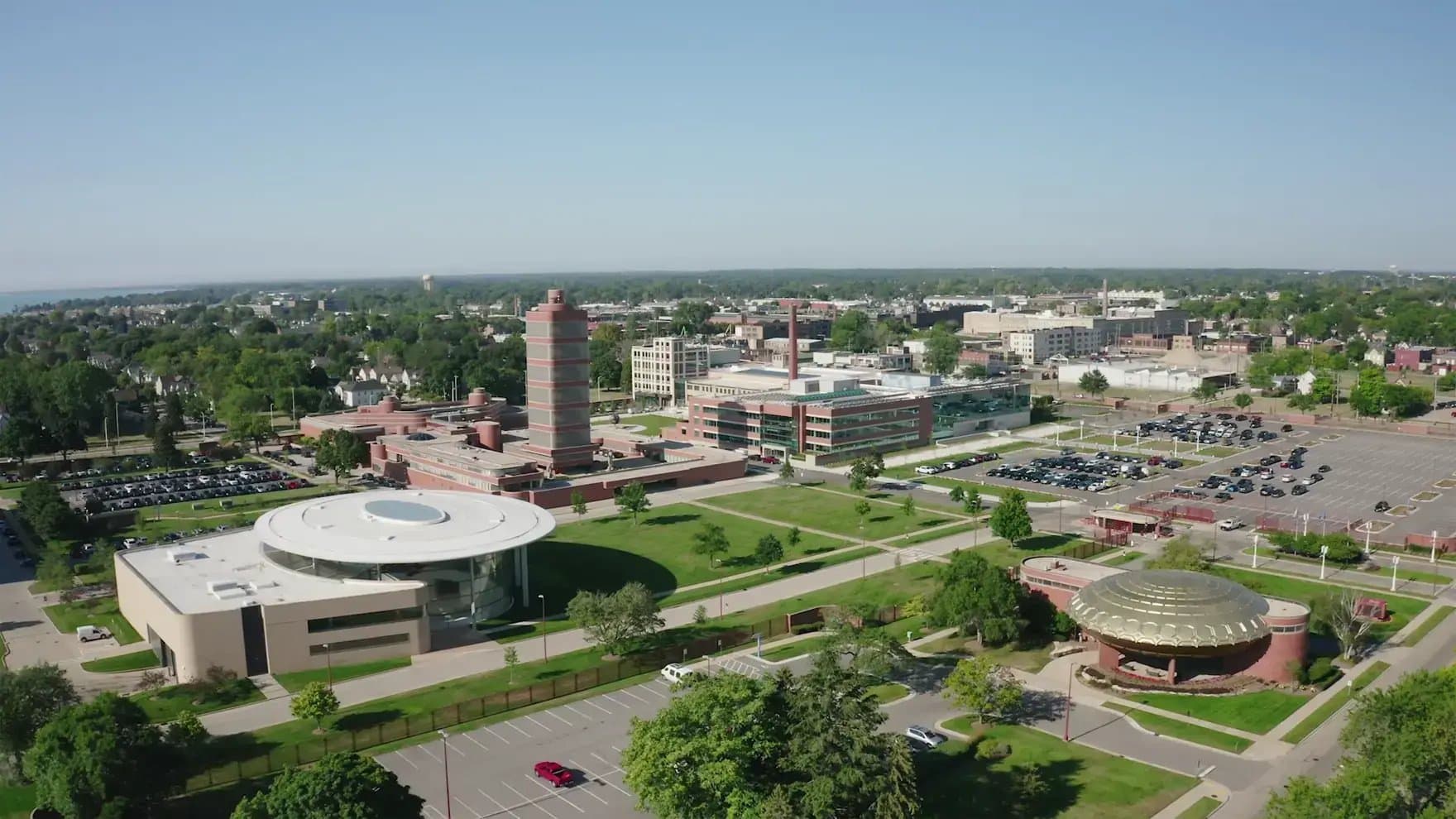 A sprawling complex of modern, circular and rectangular buildings sits on a lush green campus with surrounding parking lots. The area is bordered by a residential suburban neighborhood and distant trees under a clear blue sky.