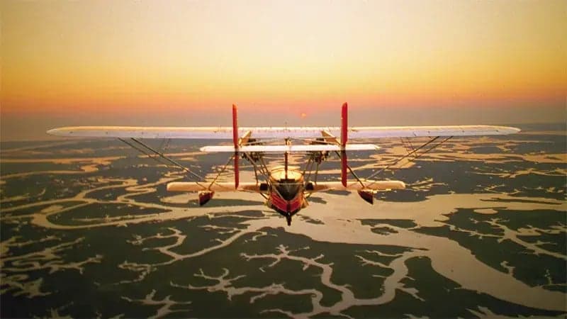 A biplane flies at sunset over a sprawling river delta, with the sun glowing on the horizon and reflecting off the water.