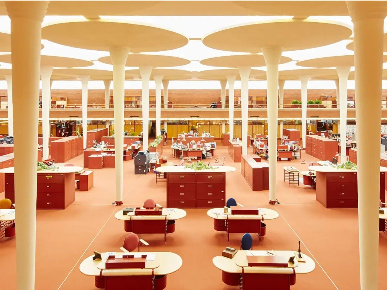 A spacious open-plan office features red desks and cabinets, arranged symmetrically. Circular skylights and white columns dominate the ceiling. The environment is modern and well-lit.