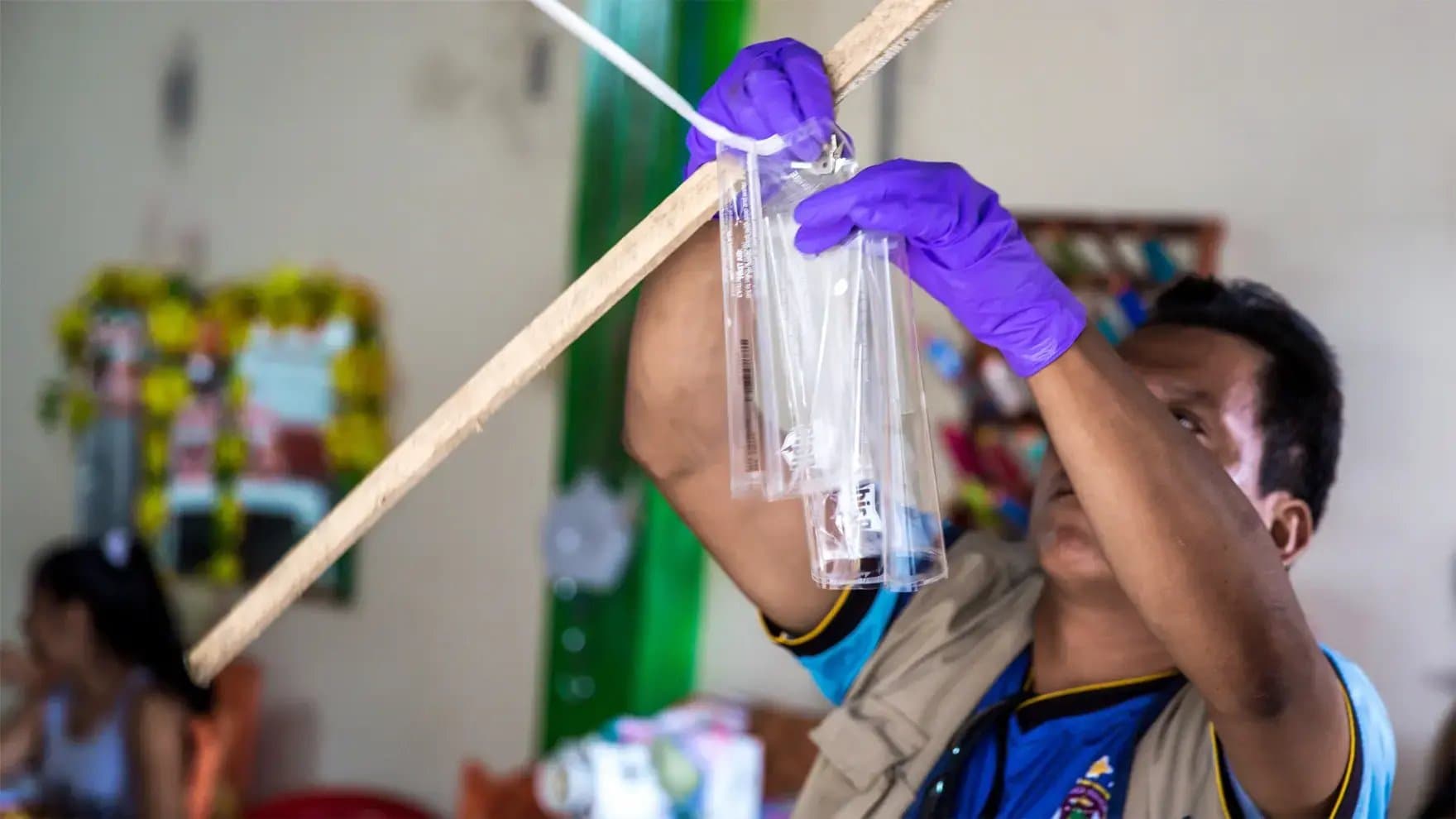 A person wearing purple gloves hangs transparent tubes on a wooden stick in an indoor setting decorated with colorful floral arrangements.