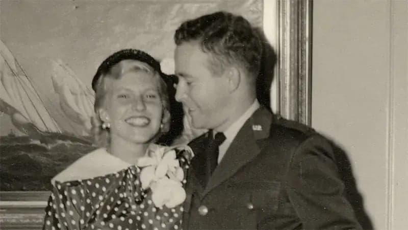 A smiling woman in a polka dot dress with a floral accessory stands next to a man in a military uniform; a sailing painting is visible in the background.