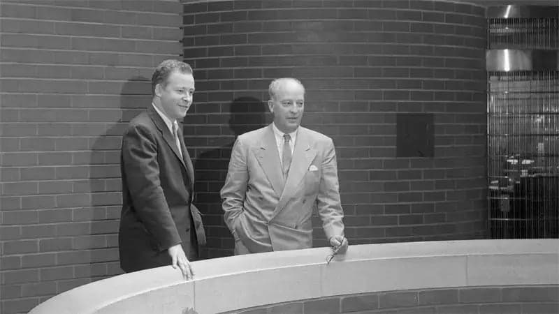 Two men stand side by side, leaning slightly against a curved railing, in front of a brick wall in a modern indoor setting.