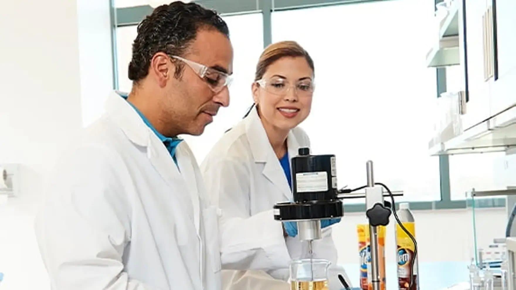 Two scientists in lab coats and safety glasses operate a laboratory device, mixing liquids in a beaker. They are in a bright, modern laboratory with shelves and equipment.
