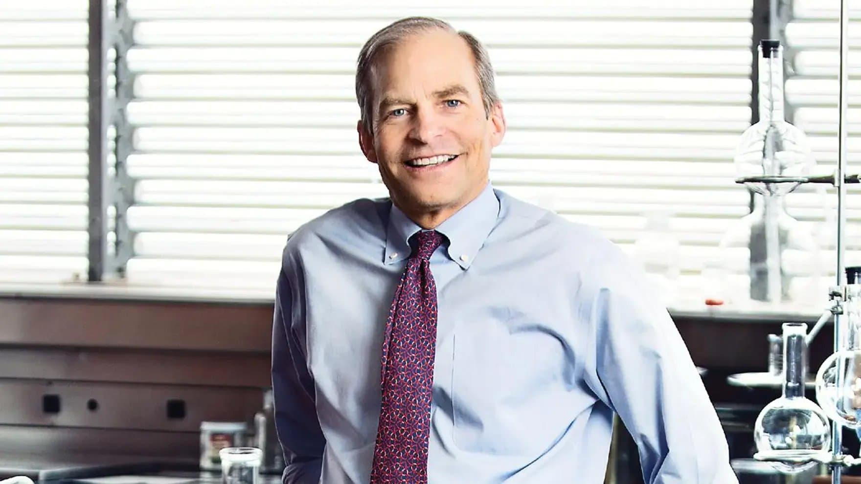 A person in a light blue shirt and red patterned tie smiles while standing in a laboratory setting, with glassware and shelves visible in the background.