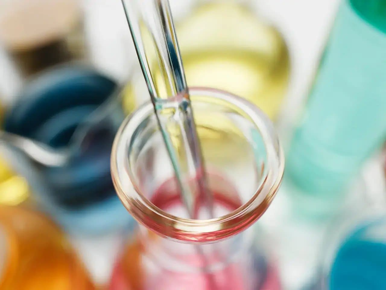 A test tube filled with pink liquid has a glass stirrer inside. Background includes blurred, colorful laboratory containers with blue, yellow, and green hues.