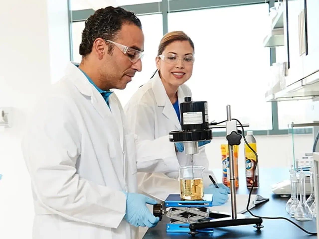 Scientists wearing lab coats and goggles are using a mixer to blend liquid in a beaker on a lab counter, surrounded by various equipment and chemicals in a bright laboratory setting.