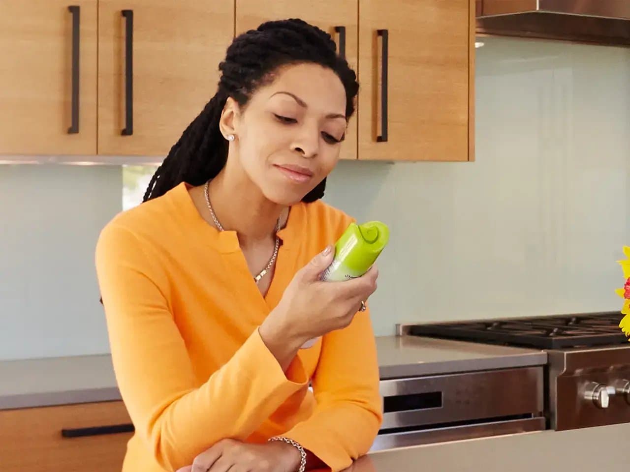 A person in an orange shirt holds and examines a green bottle, standing in a modern kitchen with wooden cabinets and stainless steel appliances.