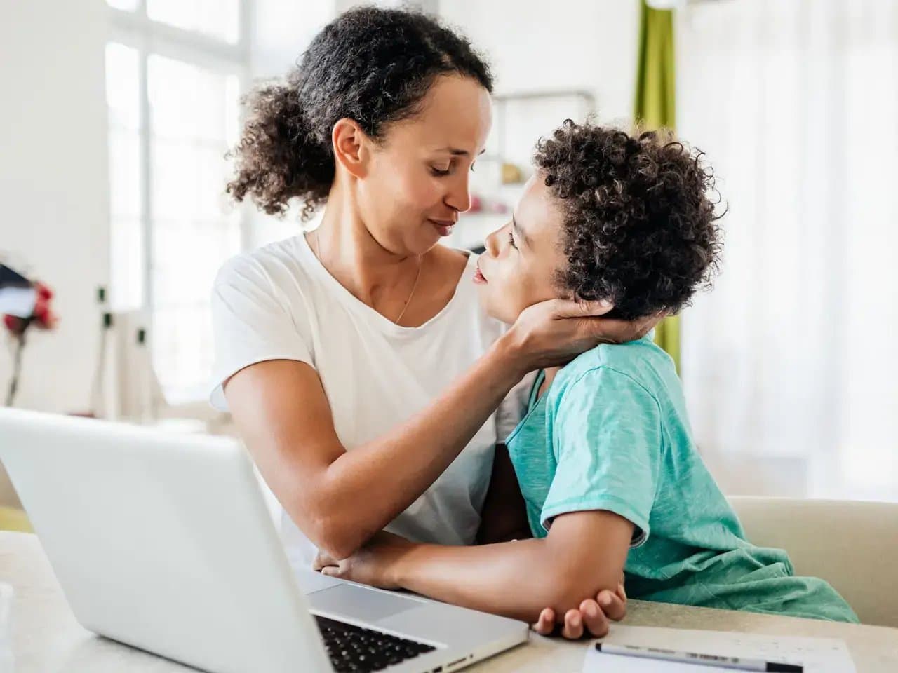 A person holds a child's face tenderly, sitting together at a table with a laptop, in a bright room with a large window and green curtains.