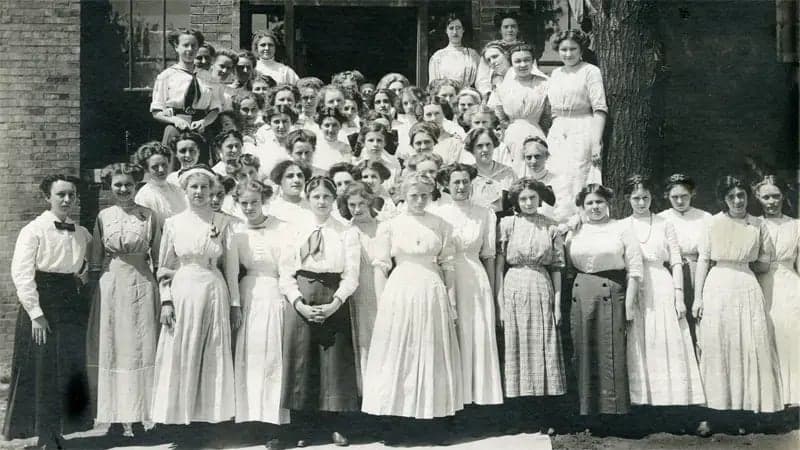 A group of women, in early 20th-century attire, stand together posing for a photograph. The background features a brick building and a large tree, indicating an outdoor setting.