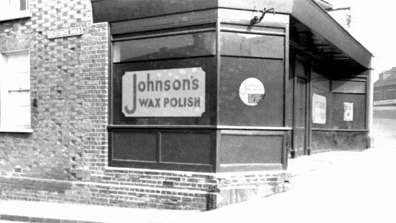 Shopfront displays a large "Johnson's Wax Polish" sign on a corner building with brick walls. Additional signage appears in windows, situated on a street. A street sign reads "Exile St."