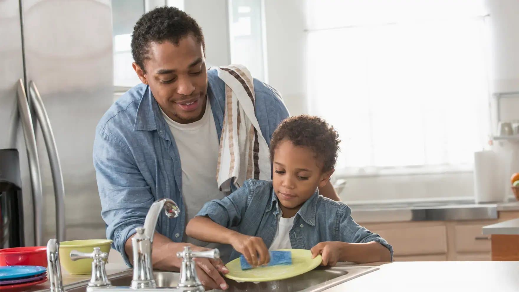 Father and son wash dishes together at a kitchen sink. The father smiles, guiding his son who uses a sponge on a plate. Sunlight filters through a window.
