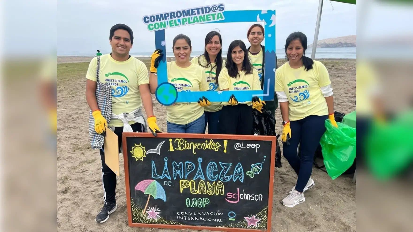 A group of six people wearing matching shirts and gloves participate in a beach cleanup, holding a frame and sign reading "COMPROMETID@S CON EL PLANETA" and "Limpieza de Playa, Conservación Internacional."