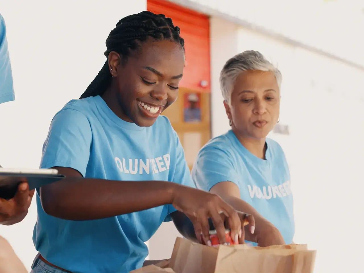 Two individuals wearing blue shirts labeled "VOLUNTEER" smile while packing items into brown paper bags on a table, situated in a brightly lit indoor setting.