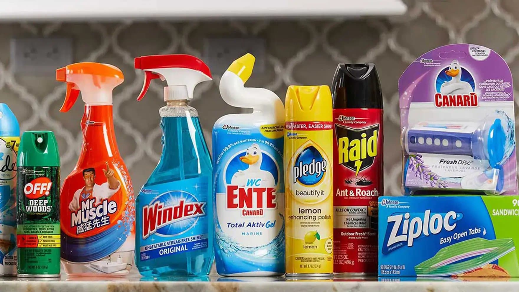 Assorted household products line up on a counter, including sprays like OFF! Deep Woods, Mr. Muscle, Windex, Pledge, Raid, and items like Ziploc bags and Canard Fresh Disc, against a patterned backsplash.