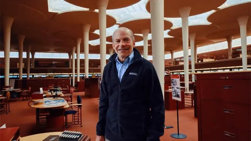 A person stands smiling, wearing a dark jacket, in an open-plan office with unique circular ceiling cutouts and tall columns, surrounded by desks and chairs.