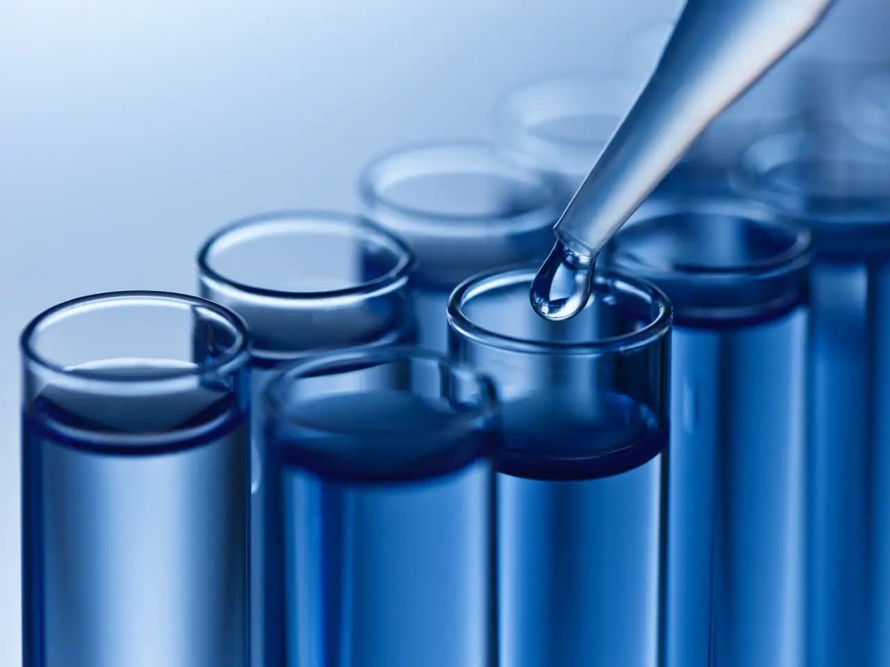 Test tubes filled with blue liquid; a pipette drips into one. The scene is a scientific laboratory setting with a cool blue tone, suggesting precision and experimentation.