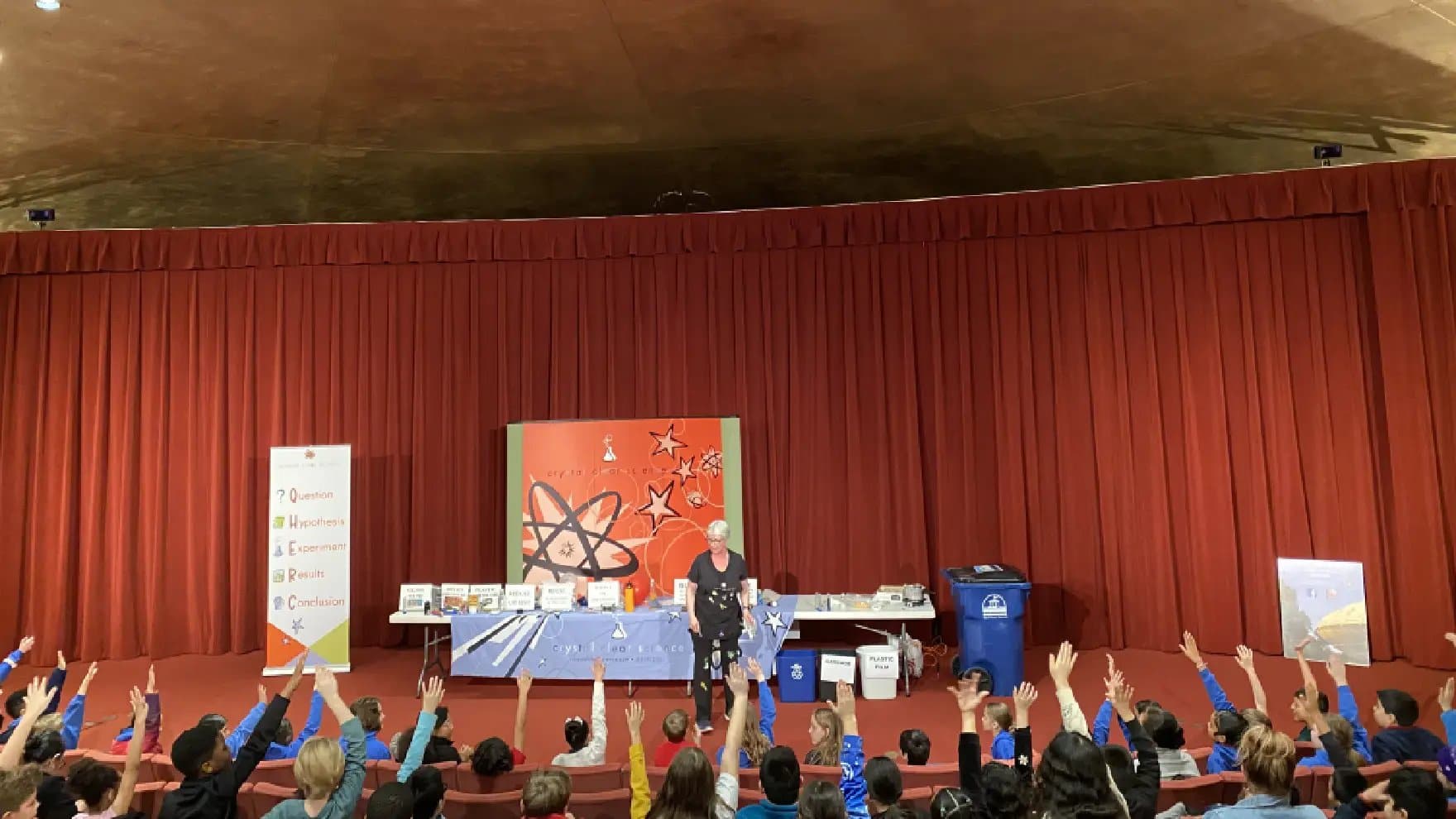 A presenter stands on stage with kids raising hands, against a red curtain backdrop. Educational displays and bins are on tables. A banner reads: "Question, Hypothesis, Experiment, Results, Conclusion."