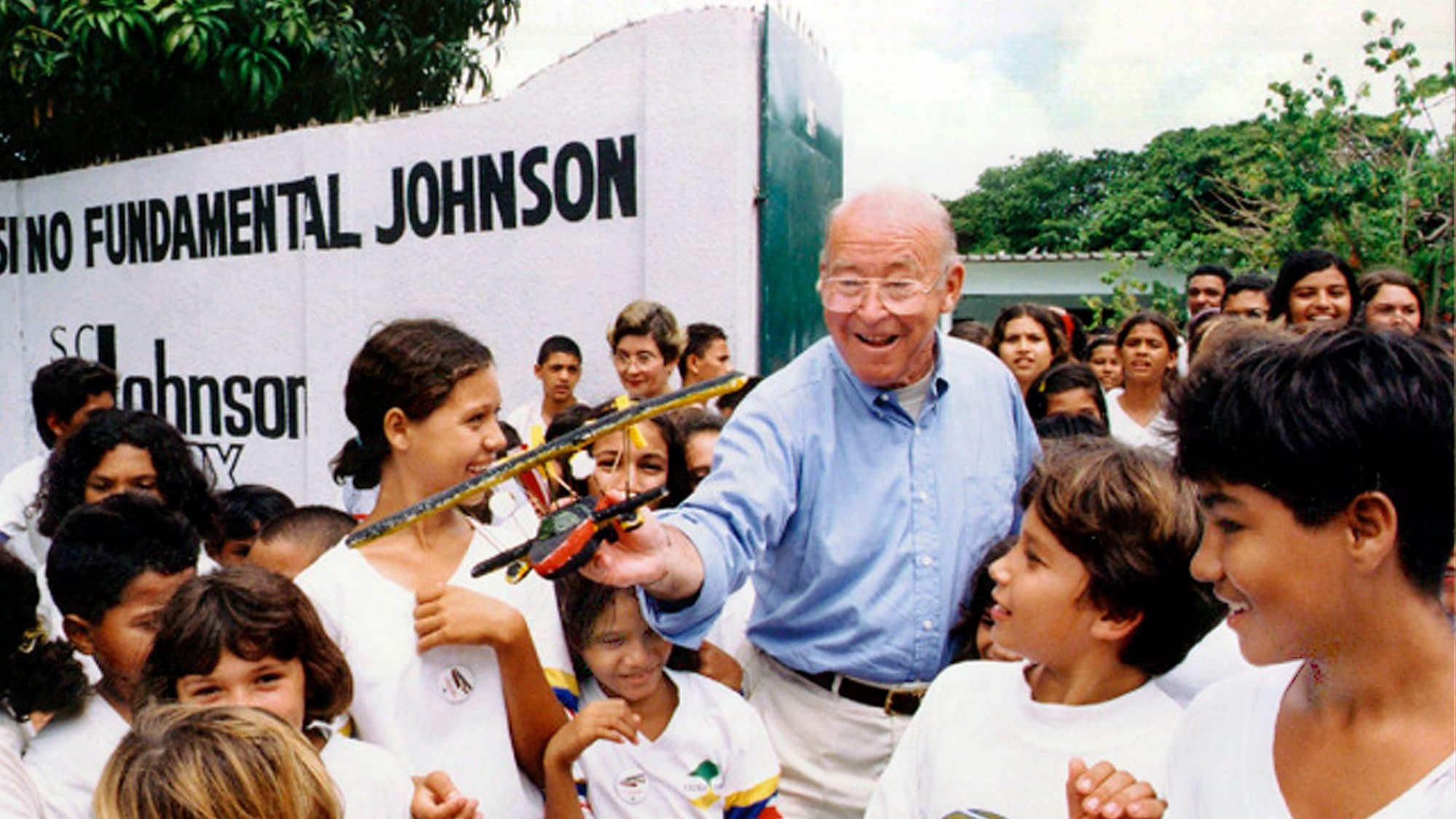 A man in a blue shirt shares a model airplane with smiling children. They stand outdoors near a wall labeled "ENSINO FUNDAMENTAL JOHNSON" and "S.C. Johnson Wax," surrounded by greenery.